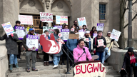 A person in a hoodie speaks into a microphone at a podium. More than a dozen people hold union signs, showing SEIU and messages like "Your tuition isn't paying your teachers."
