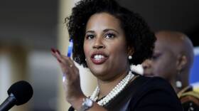 A woman wearing a pearl necklace and earrings gestures with her hand while speaking into a microphone.