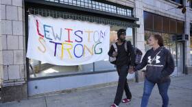 Two people hold hands and look at one another as they walk on a sidewalk past a handmade banner that says "Lewiston Strong"