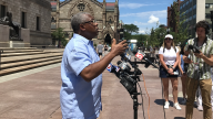 A man in a blue shirt speaks in front of a group of microphones in front of the steps of the Boston Public Library.
