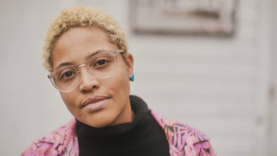 A close-up image of a person with short blonde hair, glasses, turquoise earrings and a black turtleneck.