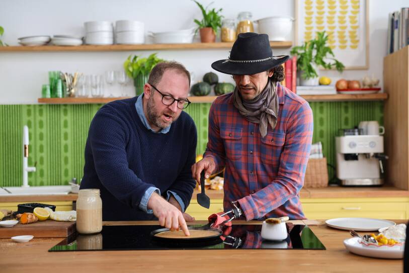 Man with glasses and beard in navy blue sweater cooks at the stovetop with a man with a prosthetic arm in a plaid shirt and hat