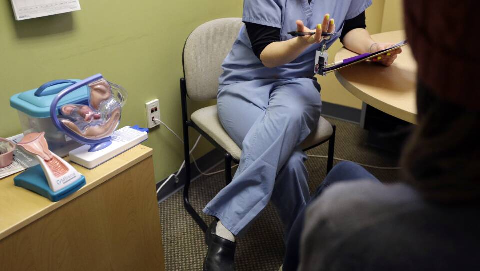 A person wearing medical scrubs sits in a chair by a table with plastic displays showing the female reproductive system.