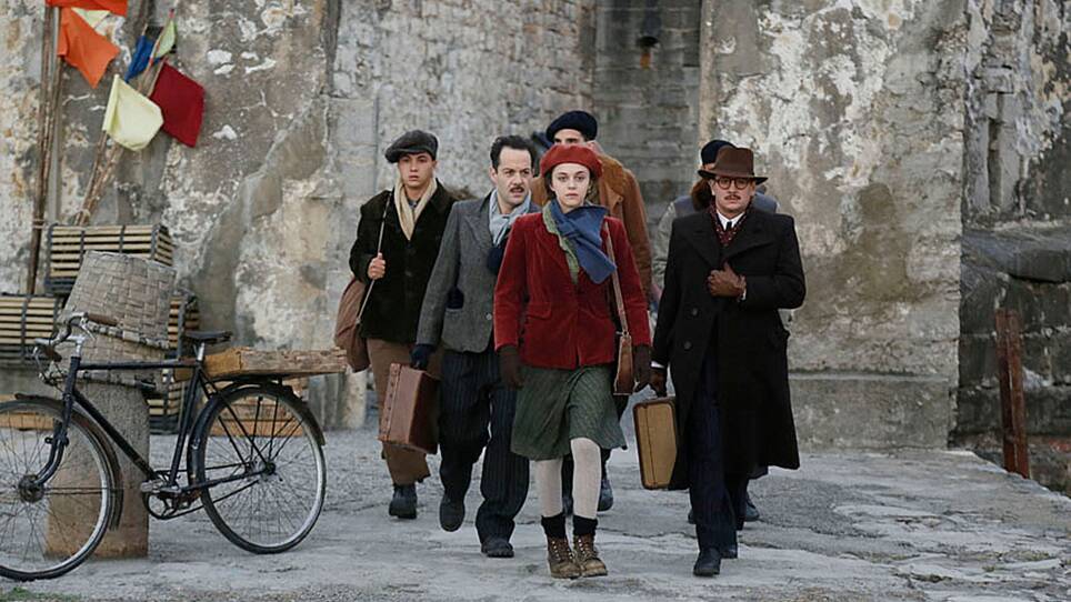 A group of men walk toward camera, led by a young woman in a red hat and coat. Behind them is a dingy stone wall.