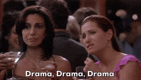 A woman holding two martinis says "drama, drama, drama" to her friend.
