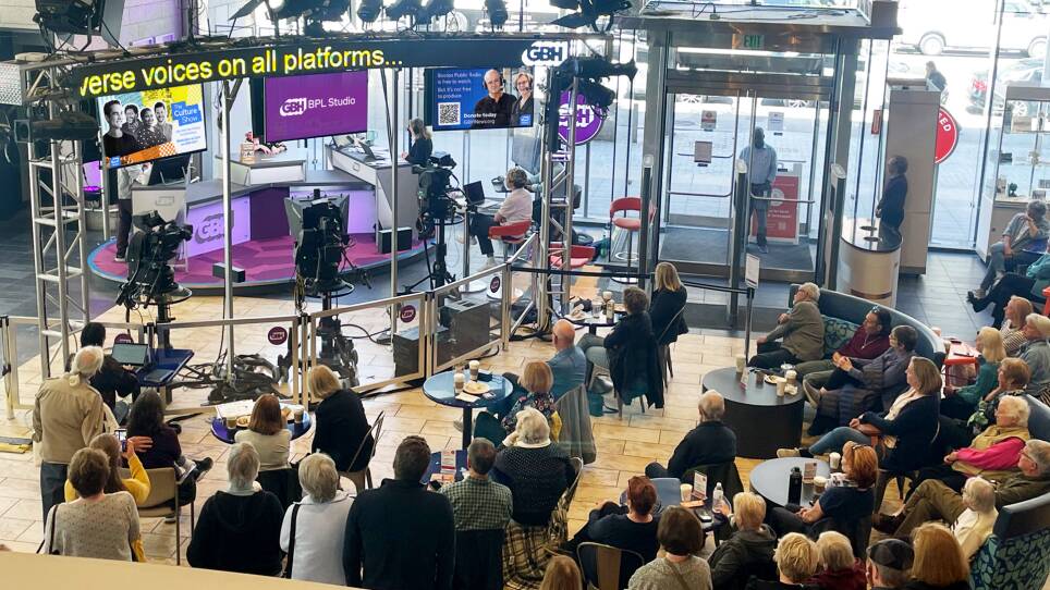 Overhead view of the GBH Boston Public Library Studio with people sitting at tables and chairs watching the broadcast of Boston Public Radio