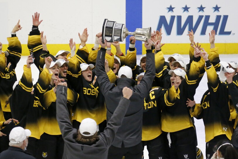 Dozens of women hockey players in black and yellow jerseys raise their hands in celebration as a coach hoists a silver trophy above his head.
