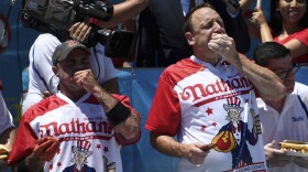 Two men in red and white jerseys with the word Nathan's shove hot dogs into their mouths.