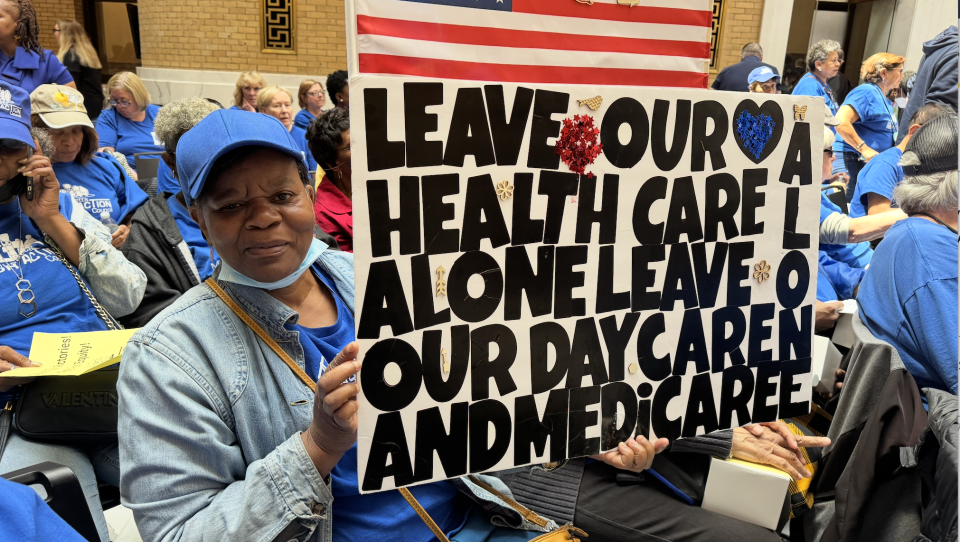A woman holds up a sign that reads 'leave our health care alone, leave our day care and Medicare alone.' She is surrounded by people wearing blue t-shirts.