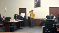 A man in a yellow sweater stands in court, his hands cuffed behind his back.