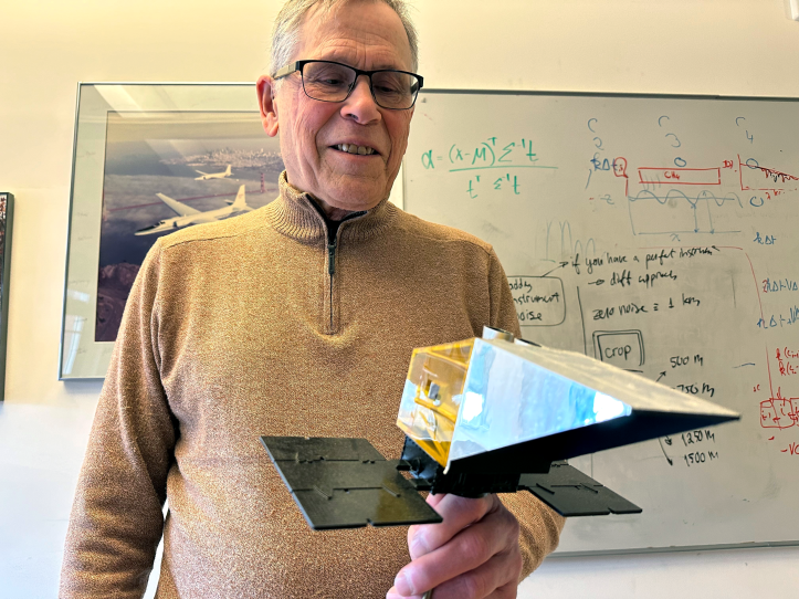 A man proudly holds a small model, with an equations-covered whiteboard in the background.