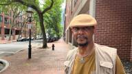 A man wearing a brimless felt cap and a cargo vest poses for a portrait standing on red brick Boston sidewalk.