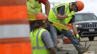 A male construction worker wearing an orange hard hat, goggles and a yellow safety vest wields a hammer in his right hand as he leans down to drive a steel spike into the ground. Two other workers on the crew are also pictured, standing among orange safety barrels typical of road construction projects.