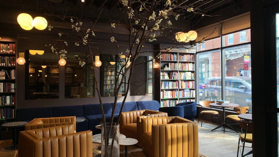 in the foreground of this lounge is a table with a vase of flowering branches, in the background is a long, blue sectional sofa, which is flanked by bookcases. In the middle of the lounge area there are two leather arm chairs facing two other leather arm chairs. Between them are small circular tables to hold books, food and beverages. To the right of the lounge is a large window that looks out onto a street. The room is lit by natural light and overhead light fixtures.