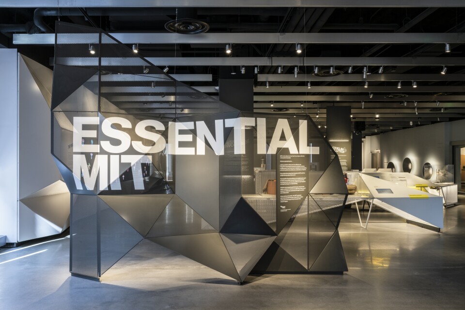 This photo is of the entrance to the new MIT Museum, which emphasizes advances in technology. The floors are poured concrete. There is a large grey screen made out of triangles, which fold and bend as if it were origami. Across the screen in white all capital letters it says "ESSENTIAL MIT." Beyond the screen you can see white museum display cases in the background
