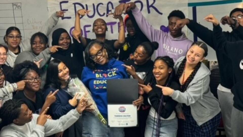 Tanisha Milton, 8th-grade teacher at TechBoston Academy and one of BPS's Educators of the Year, surrounded by 16 students pointing at her. Milton is wearing a blue shirt and glasses and holding an award in front of a sign that says "Educator of the Year."