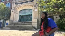 A teenager sits on a block by the sidewalk outside a large school building.