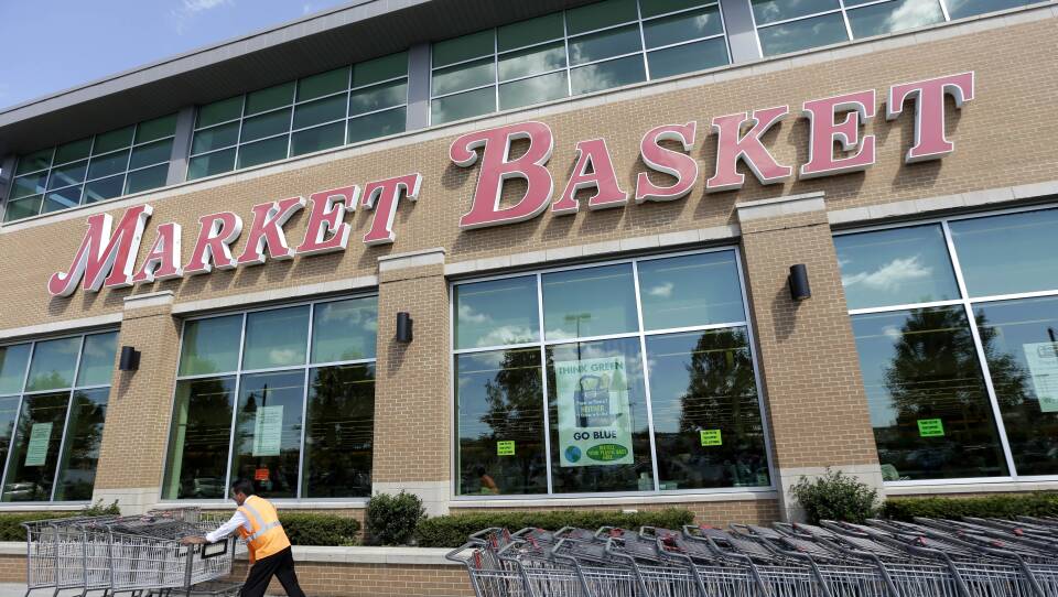A worker pushes a line of grocery baskets in front of a large store front with the Market Basket logo above.