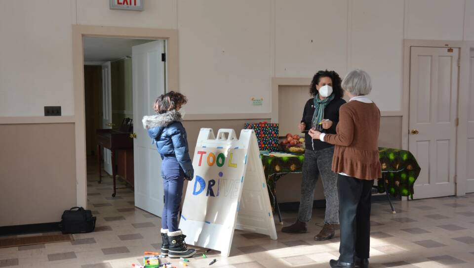 Three people stand in the lobby of a church that will now be a community center for people of color. A child stands in front of a sign that says "Tool Drive" in rainbow colors, two older women stand and talk to each other in front of a table with food on it.
