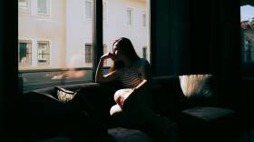 A woman puts a hand up to her face as she looks out a window. She's sitting in a dark apartment.