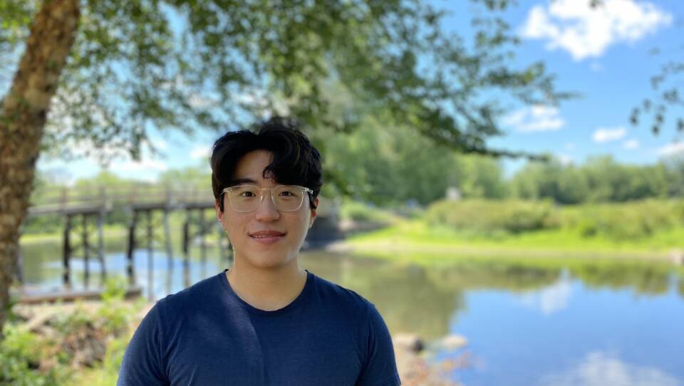 A 21-year-old Korean-American man in a dark blue T-shirt and transparent frames on his glasses looks at the camera and grins. He's standing in front of a body of water with a footbridge going over it.