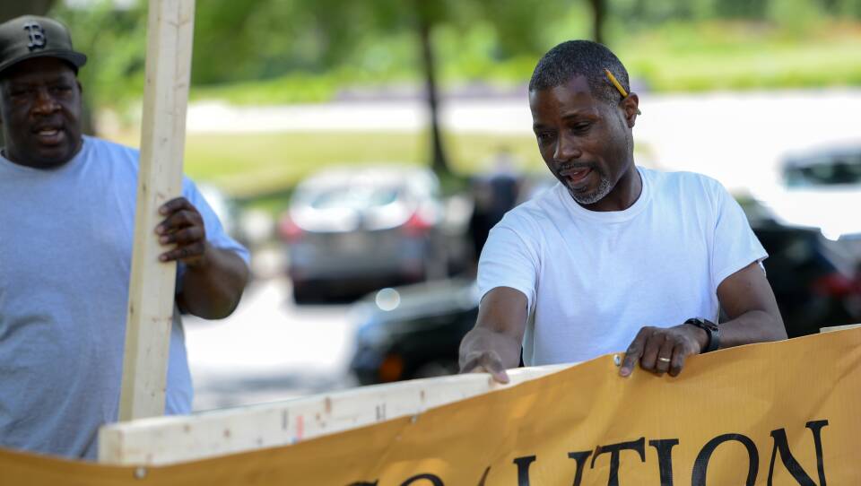 A man holds a black and gold banner against a 2 by 4 piece of lumber as another man approaches with a second board to help assemble a frame for sign.