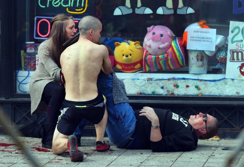 A shirtless Wheeler and a woman kneel next to a man lying on the ground in pain. He's applying pressure to an elevated leg that's blocked by Wheeler's body. Splatters of blood are on the ground.