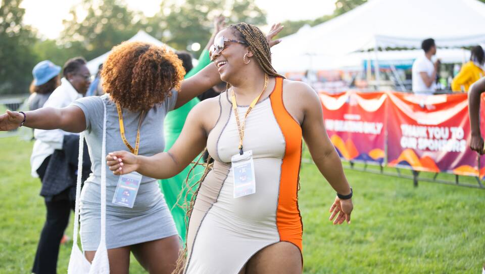 Two women dance in the middle of a grassy area. One smiles widely wearing sunglasses.