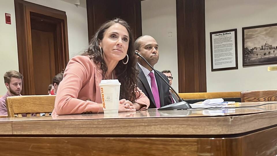 A woman with curly brown hair, wearing a pink jacket, sits at a wooden table with a microphone and a Dunkin' Donuts cup in front of her.