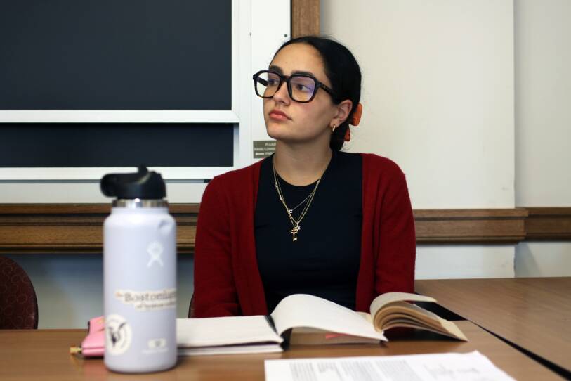 A woman in black glasses looks to her right as a book and notebook lay open on a table in front of her.