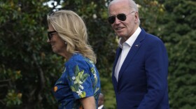 A woman in a blue floral dress and a man in a navy blue suit, both wearing sunglasses, walk outdoors.