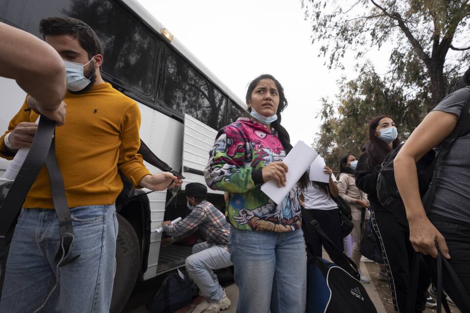 A woman in a graffiti-style sweatshirt looks confused as she holds a sheet of paper and a bag, just after getting off a large bus with a crowd of other people.