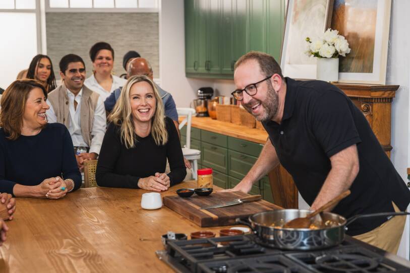 Man with a beard and glasses in a black polo shirt stands at a kitchen stovetop while an audience looks on