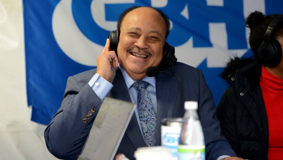 A man smiles as he listens to sound coming through a radio headset, holding one hand up to an earpiece to hold it closer to his head.