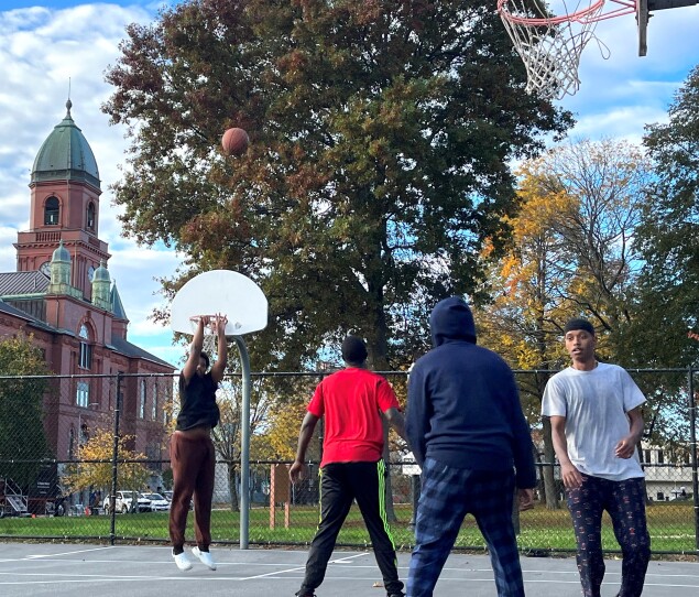 Four young men play basketball on an outdoor court with autumn leaves and Lewiston City Hall in the background.
