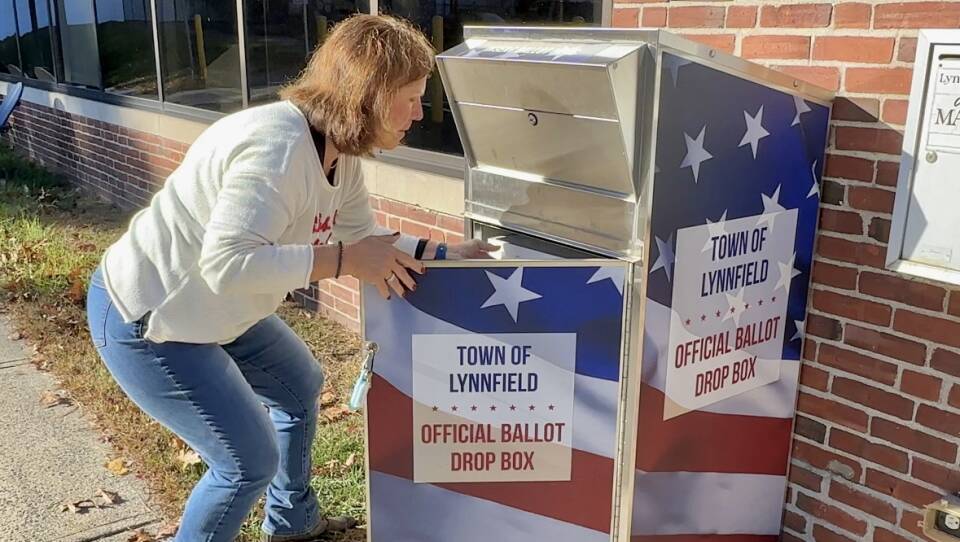 A town employee, a woman in jeans and a white sweater, bends toward a ballot drop box that she has opened to collect ballots for the town of Lynnfield.
