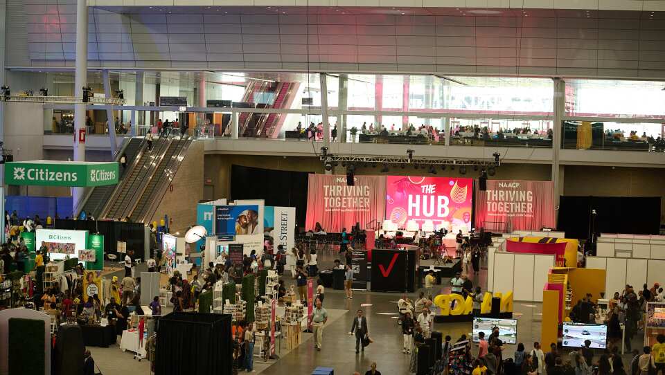 A crowded convention hall features booths and a lit stage at the base of escalators.