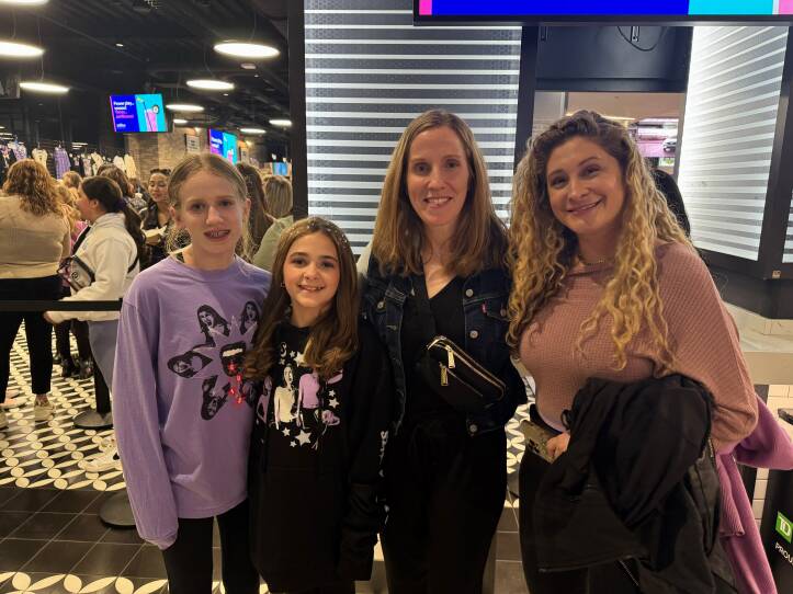 Two young girls and two adult woman stand inside TD Garden concert arena. The girls are wearing merchandise for singer Olivia Rodrigo.