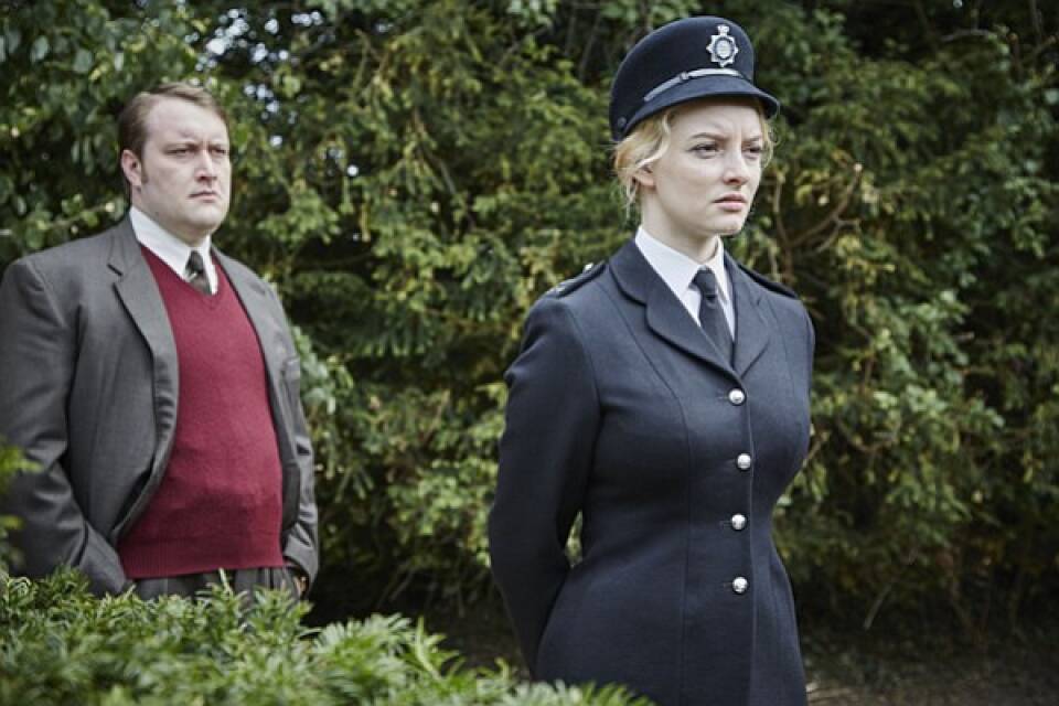 In a verdant garden, a young policewoman in a vintage uniform stares off into the distance. Just behind her, a man in a suit with a maroon sweater vest looks in the same direction.