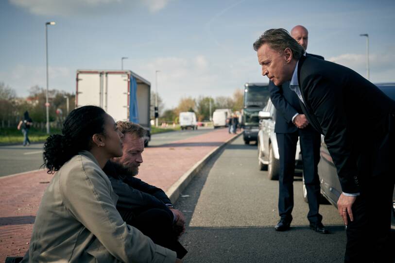 A man and a woman sit on the island in the middle of a street, facing the right of the frame. On either wide of the street, cars are stopped. To the right of the frame, a man in a suit bends over, hands on his knees, talking to the man and woman sitting. In the background of the photo, a bodyguard stands at ease.