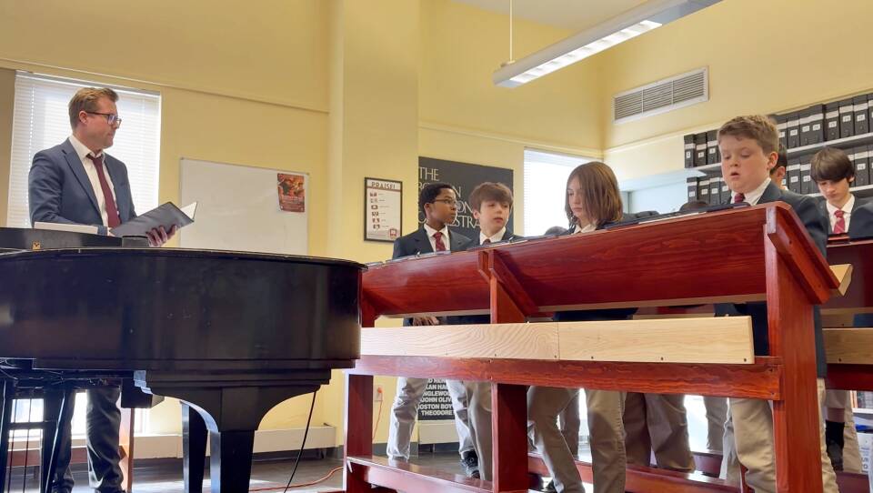 A man stands with a folder open as he speaks to a group of boys standing behind music stands.