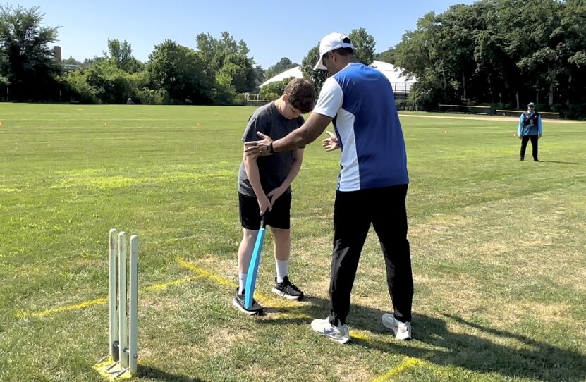 A young boy in a gray tshirt and shorts looks down at a bright blue plastic cricket bat he's holding. A man in a blue and white tshirt stands in front of the boy holding his arms out to demonstrate good batting stance.