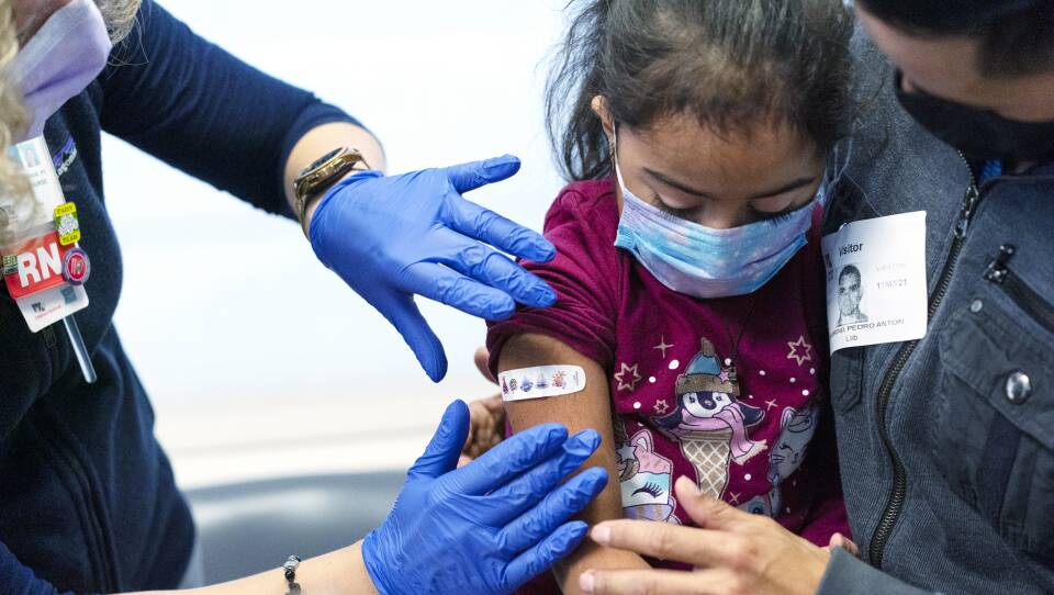 A nurse wearing gloves places a bandage on the arm of a young girl as she sits on her father's lap.
