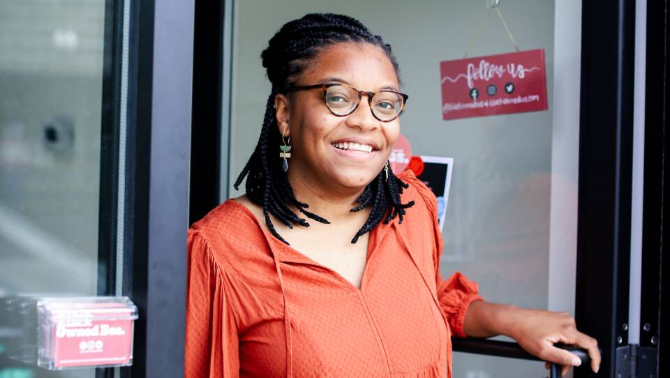 A Black woman in an orange blouse holds open the door of a shop.