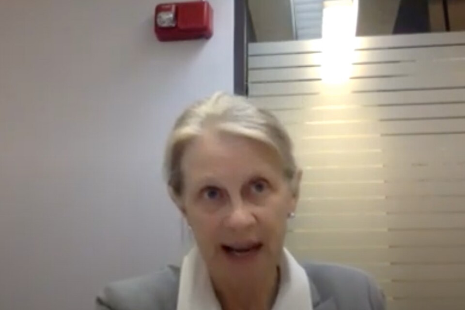 A woman in a gray blazer is mid-speech.