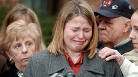 A young woman cries as she stands in the middle of a group of people. One person has their hand resting on her shoulder to console her.