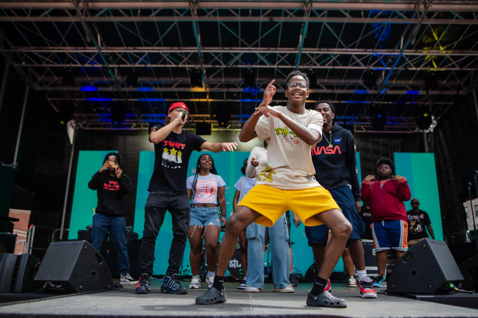 A young man in a white t-shirt and yellow shorts dances in the foreground with other young people performing and smiling behind him.