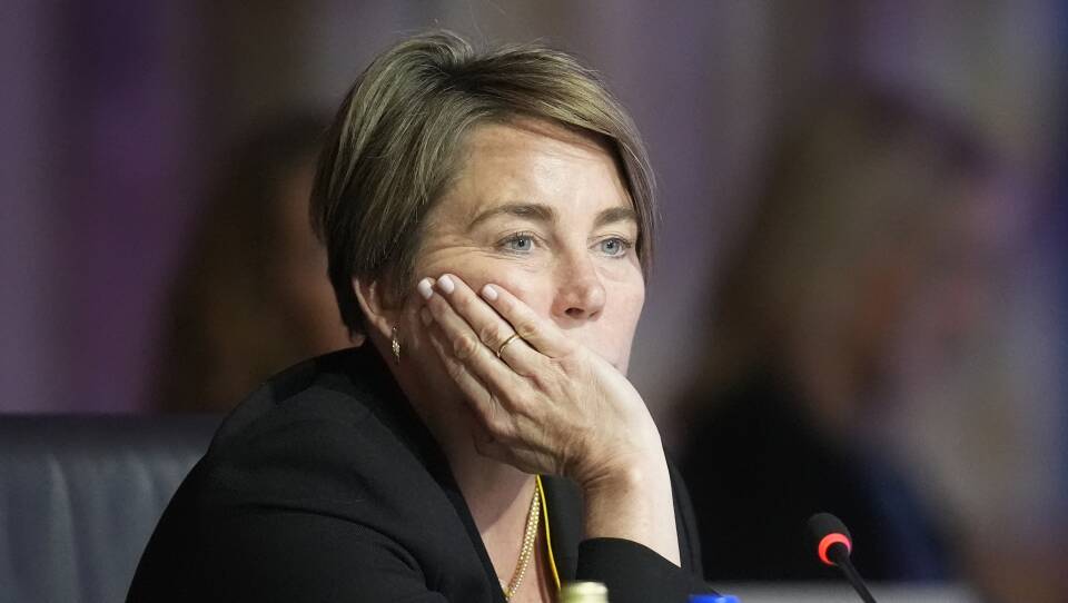 A woman wearing a black blazer rests her face in her right hand and looks forward in front of a small microphone.