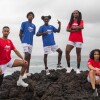 Five athletes in shorts and red and blue t-shirts pose on beachfront rocks with the ocean behind them.
