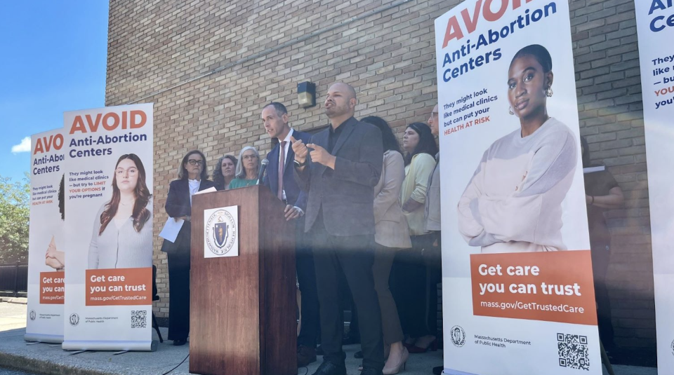 A group of people stand at a podium next to banners that say 'avoid anti-abortion centers.'